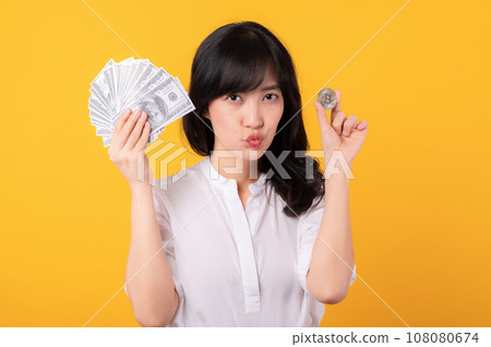 Portrait beautiful young asian woman enterpriser with doubt face wearing white shirt and red plants holding cash dollar money and crypto digital currency isolation on yellow background. Wealth concept Portrait beautiful young asian woman enterpriser with doubt face wearing white shirt and red plants holding cash dollar money and crypto digital currency isolation on yellow background. Wealth concept 108080674