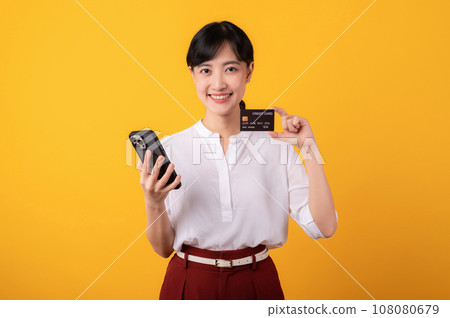 Portrait beautiful young asian woman enterpriser happy smile wearing white shirt and red plants holding credit card and smartphone isolation on yellow background. Online shopping concept. 108080679