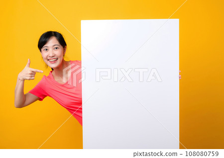 young asian sports fitness woman happy smile wearing pink sportswear standing behind the white blank banner or empty space advertisement board against yellow background. wellbeing lifestyle concept. 108080759