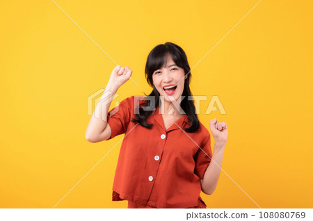 Portrait young beautiful asian woman happy smile dressed in orange clothes showing fist up hand gesture isolated on yellow studio background. Successful hooray celebrate young person concept. Portrait young beautiful asian woman happy smile dressed in orange clothes showing fist up hand gesture isolated on yellow studio background. Successful hooray celebrate young person concept. 108080769