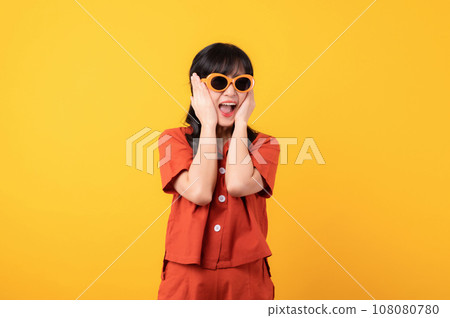 Portrait young beautiful asian woman happy smile dressed in orange clothes and sunglasses looking surprised, reacting amazed, raising eyebrows impressed isolated on yellow studio background. 108080780