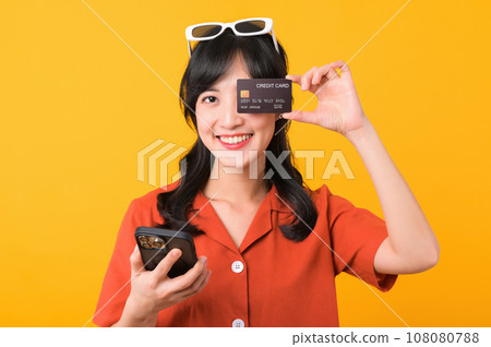 Portrait young beautiful asian woman happy smile dressed in orange clothes using smartphone and showing credit card isolated on yellow background. Pay and purchase shopping payment concept. Portrait young beautiful asian woman happy smile dressed in orange clothes using smartphone and showing credit card isolated on yellow background. Pay and purchase shopping payment concept. 108080788