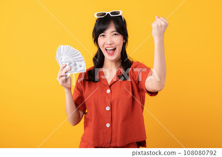 Portrait young beautiful asian woman happy smile dressed in orange clothes holding cash money and celebrate her success isolated on yellow background. Be rich concept. Portrait young beautiful asian woman happy smile dressed in orange clothes holding cash money and celebrate her success isolated on yellow background. Be rich concept. 108080792
