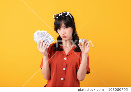 Portrait young beautiful asian woman happy smile dressed in orange clothes holding cash money and crypto bitcoin digital currency celebrate her success isolated on yellow background. Be rich concept. 108080793