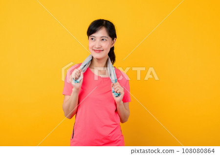 Portrait young asian sports fitness woman happy smile wearing pink sportswear and face towel doing exercise training workout against yellow studio background. wellbeing and healthy lifestyle concept. 108080864