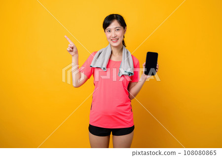 Portrait young asian sports fitness woman happy smile wearing pink sportswear and smartphone doing exercise training workout against yellow studio background. technology wellness lifestyle concept. 108080865