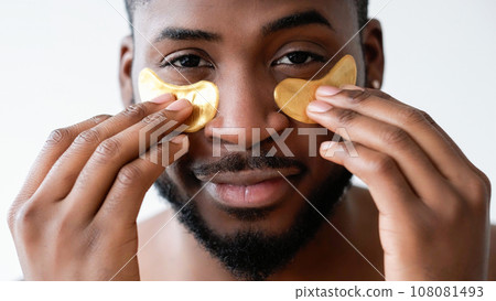 Man facial care. Skin treatment. Anti aging procedure. Confident satisfied guy applying gold hydrogel eye patches smiling isolated on white background. 108081493