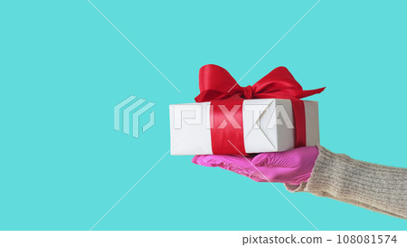 Birthday gift. Offering present. Unrecognizable human hand in protective gloves holding wrapped box hygiene isolated on blue empty space background. Birthday gift. Offering present. Unrecognizable human hand in protective gloves holding wrapped box hygiene isolated on blue empty space background. 108081574
