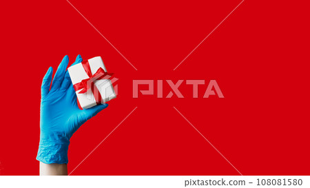 Present delivery. Epidemic gift. Unrecognizable human hand in protective glove holding wrapped box quarantine hygiene measures isolated on red empty space background. 108081580