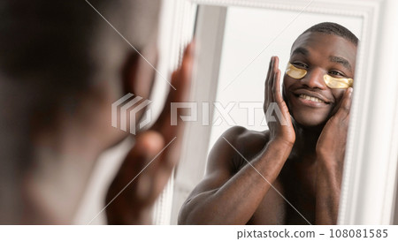 Metrosexual skincare. Morning fun. Relaxed confident smiling shirtless man with gold hydrogel eye patches in white mirror satisfied with face skin. Metrosexual skincare. Morning fun. Relaxed confident smiling shirtless man with gold hydrogel eye patches in white mirror satisfied with face skin. 108081585