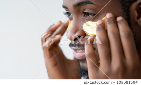 Man cosmetic. Wrinkles product. Confident guy enjoying facial skincare applying gold hydrogel eye patches on face skin isolated on white background empty space. Man cosmetic. Wrinkles product. Confident guy enjoying facial skincare applying gold hydrogel eye patches on face skin isolated on white background empty space. 108081594
