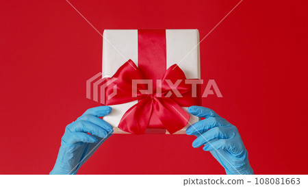Christmas gift. Pandemic New Year. Female hands in blue medical gloves holding box with present isolated on red copy space background. Christmas gift. Pandemic New Year. Female hands in blue medical gloves holding box with present isolated on red copy space background. 108081663
