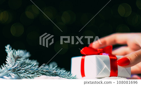 Christmas gift. Holiday surprise. Greeting congratulation. Woman hand putting present box taped red ribbon on snowy pine branch decoration bokeh green lights background copy space. 108081696