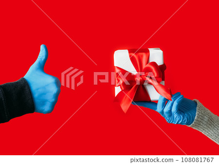 Gift delivery. Present offering. Unrecognizable human hands in protective gloves holding wrapped box thumb up like gesture isolated on red background. 108081767