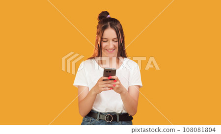 Mobile chatting. Text message. Happy pleased smiling woman typing in modern smartphone social media application isolated on orange background copy space. 108081804