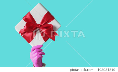 Gift delivery. Virus present. Unrecognizable human hand in protective glove holding wrapped box quarantine hygiene measures isolated on blue empty space background. Gift delivery. Virus present. Unrecognizable human hand in protective glove holding wrapped box quarantine hygiene measures isolated on blue empty space background. 108081840