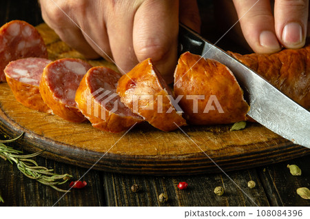 Close-up of the chef hands with a knife cut meat sausage on a cutting wooden board. Concept of cooking fast food or sandwich 108084396