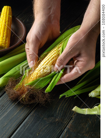 Peeling heads of corn from the shell by the hands of the cook before grilling. Work environment on the kitchen table. Copy space. Peeling heads of corn from the shell by the hands of the cook before grilling. Work environment on the kitchen table. Copy space. 108084400