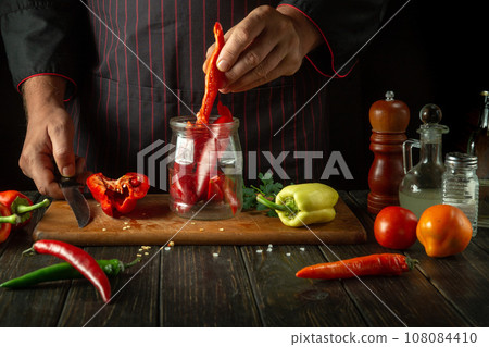 Preserving capsicum annuum in a spice jar. A cook puts red bell pepper into a jar in a restaurant kitchen. Concept of preparing or canning vegetables 108084410