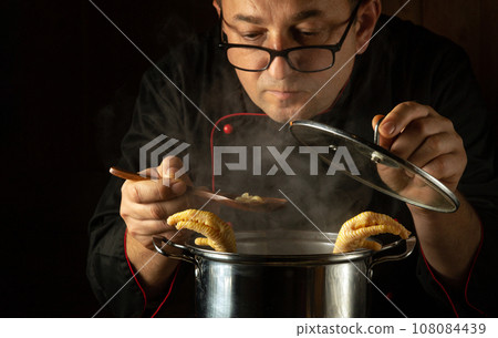 The cook prepares a delicious soup with pasta and chicken in a saucepan. Spoon in the chef's hand. Checking food readiness The cook prepares a delicious soup with pasta and chicken in a saucepan. Spoon in the chef's hand. Checking food readiness 108084439