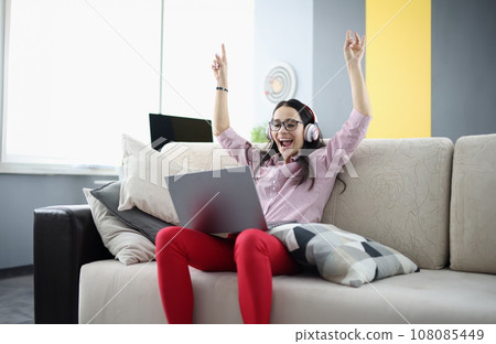 Rejoicing woman is sitting on sofa with in headphones with laptop. Rejoicing woman is sitting on sofa with in headphones with laptop. 108085449