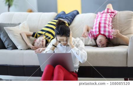 Woman sits on floor with a laptop and covers her ears with her hands. behind her on couch are children playing around. 108085520