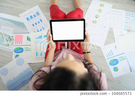 Woman is sitting on floor around documents with charts with commercial indicators holding tablet in her hands. Woman is sitting on floor around documents with charts with commercial indicators holding tablet in her hands. 108085571