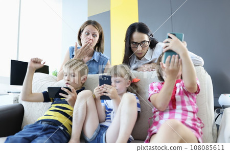 Three children are sitting on the couch with smartphones in their hands, playing online games from back of couch, two women are standing and looking frightened at phone screen. Three children are sitting on the couch with smartphones in their hands, playing online games from back of couch, two women are standing and looking frightened at phone screen. 108085611