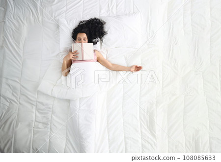 Woman lies on white bed with closed eyes book lies on her face. 108085633