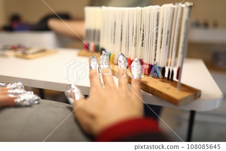 Woman's hand with foil on nails is standing next to samples of varnish with color palette. Woman's hand with foil on nails is standing next to samples of varnish with color palette. 108085645