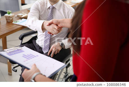 In office, man in wheelchair greets a colleague with handshake. 108086346