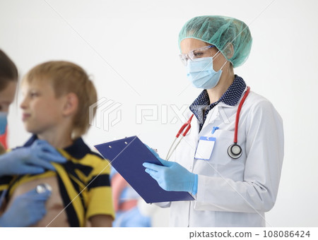 Doctor in protective medical mask writes readings on clipboard next to doctor listens with stethoscope to boy's breath. Doctor in protective medical mask writes readings on clipboard next to doctor listens with stethoscope to boy's breath. 108086424