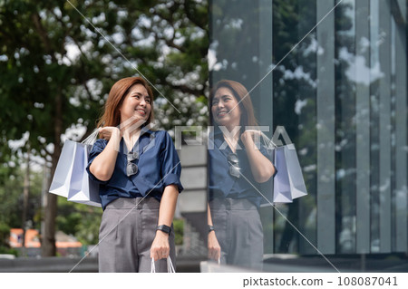 Happy beautiful young stylish woman asian with shopping bag while walking near mall on holiday Black Friday Happy beautiful young stylish woman asian with shopping bag while walking near mall on holiday Black Friday 108087041