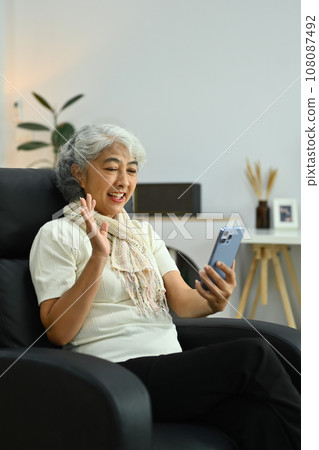 Beautiful gray haired mature woman waving hand while talking online on smartphone Beautiful gray haired mature woman waving hand while talking online on smartphone 108087492