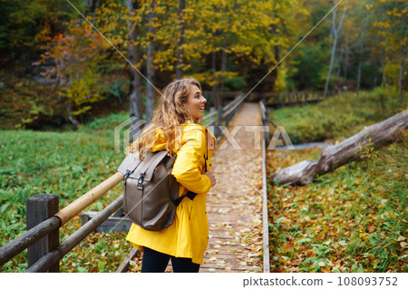 Woman in a yellow coat with backpack walks along wooden staircase, enjoying autumn scenery in forest 108093752