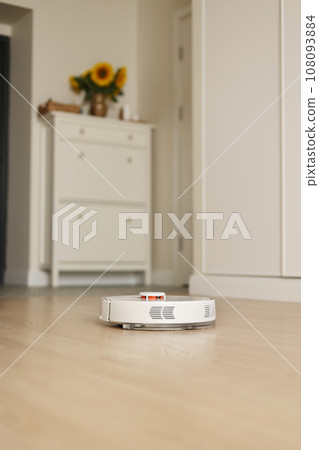 Modern wireless autonomous robotic vacuum cleaner washes wooden floor at light living room. Smart cleaning technology. Self-propelled white cleaning robot. Household appliances with remote control Modern wireless autonomous robotic vacuum cleaner washes wooden floor at light living room. Smart cleaning technology. Self-propelled white cleaning robot. Household appliances with remote control 108093884