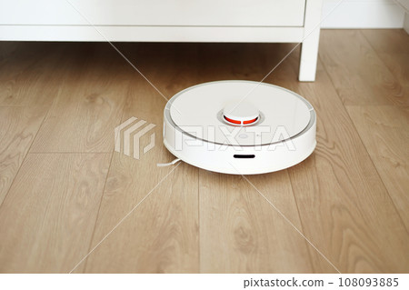 Wireless autonomous robotic vacuum cleaner vacuums with brushes under commode at living room. Self-propelled cleaning robot. Household remote control appliances. Smart cleaning technology. Wireless autonomous robotic vacuum cleaner vacuums with brushes under commode at living room. Self-propelled cleaning robot. Household remote control appliances. Smart cleaning technology. 108093885