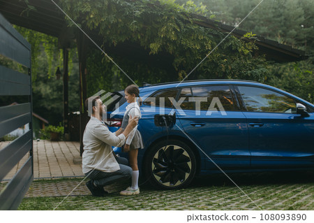 Portrait of father and daughter talking, while charging family electric car in front of house. Electric vehicle with charger in charging port. 108093890