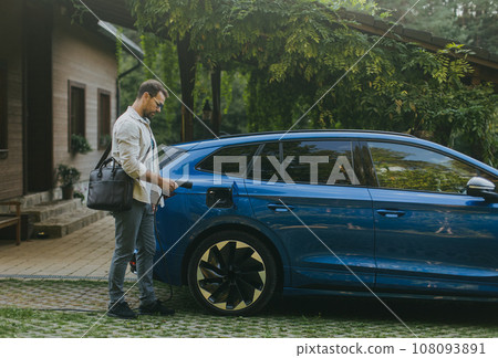 Portrait of businessman charging his electric car in front of house. Pluging charger in charging port. 108093891