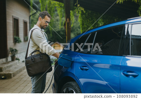 Portrait of businessman charging his electric car in front of house. Pluging charger in charging port. 108093892