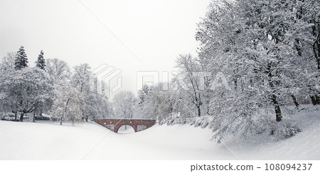 A small brick bridge over a snow-covered moat in the Zamosc city park. Snow-covered trees, snowfall. Winter landscape. 108094237