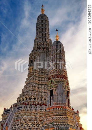The majestic Wat Arun temple in Bangkok, Thailand. It's a tall, ornate structure with multiple tiers and spires, adorned with intricate patterns and statues. The sky is blue with wispy clouds. The majestic Wat Arun temple in Bangkok, Thailand. It's a tall, ornate structure with multiple tiers and spires, adorned with intricate patterns and statues. The sky is blue with wispy clouds. 108094263