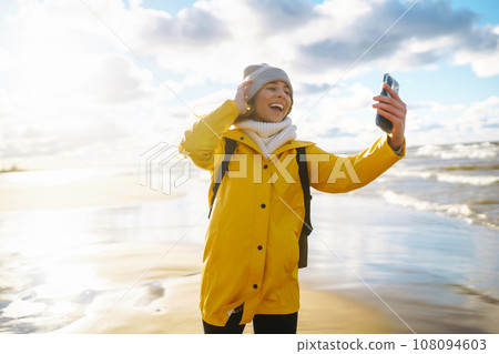 Tourist with phone. Selfie time. Tourist in yellow jacket posing by sea at sunset. Travelling. 108094603