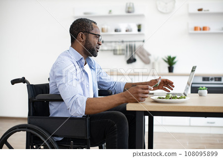 African bearded male in wheelchair using computer during mealtime in kitchen 108095899