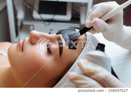 cosmetologist applying black mask on pretty woman face wearing black gloves, gorgeous woman in spa having facial procedures cosmetologist applying black mask on pretty woman face wearing black gloves, gorgeous woman in spa having facial procedures 108097027