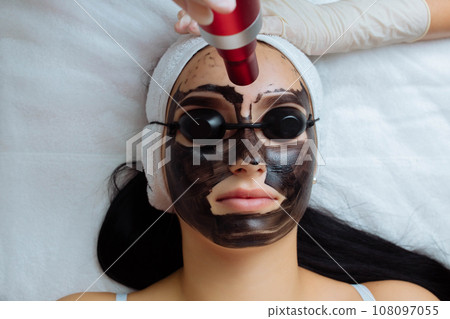 Close-up of cosmetologist's hand making hardware carbon peeling by young pretty customer woman in safety glasses headdress lying on couch in beauty salon. Facial cleaning concept. Cosmetology Services Close-up of cosmetologist's hand making hardware carbon peeling by young pretty customer woman in safety glasses headdress lying on couch in beauty salon. Facial cleaning concept. Cosmetology Services 108097055
