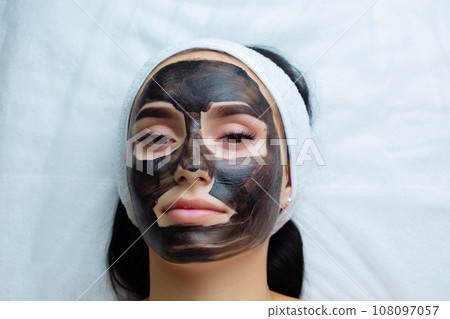 cosmetologist applying black mask on pretty woman face wearing black gloves, gorgeous woman in spa having facial procedures cosmetologist applying black mask on pretty woman face wearing black gloves, gorgeous woman in spa having facial procedures 108097057