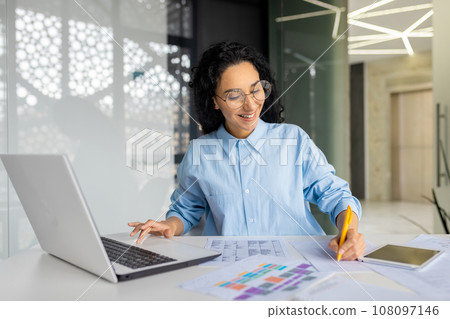 Young successful Indian woman paperwork, businesswoman records data, fills tables and checks invoices, reports and contracts. Financier satisfied results of achievement at workplace inside office. 108097146