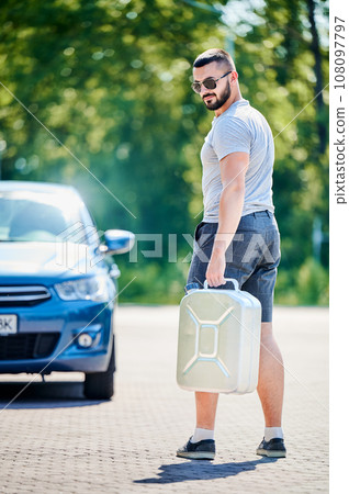 Young male looking for petrol station to fill canister. Man trying to find nearest gas station. Adult man in casual clothes holding canister on background of blue car. 108097797