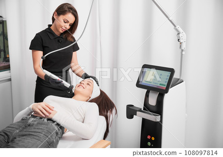 Doctor cosmetologist using radiofrequency microneedling device while performing face lifting procedure in beauty salon. Woman lying near cosmetology equipment during skincare procedure. 108097814